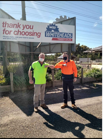 Two men standing in front of the Whiteheads timber sign, wearing high visibility shirts, work trousers and a mask. They are giving an elbow bump to each other