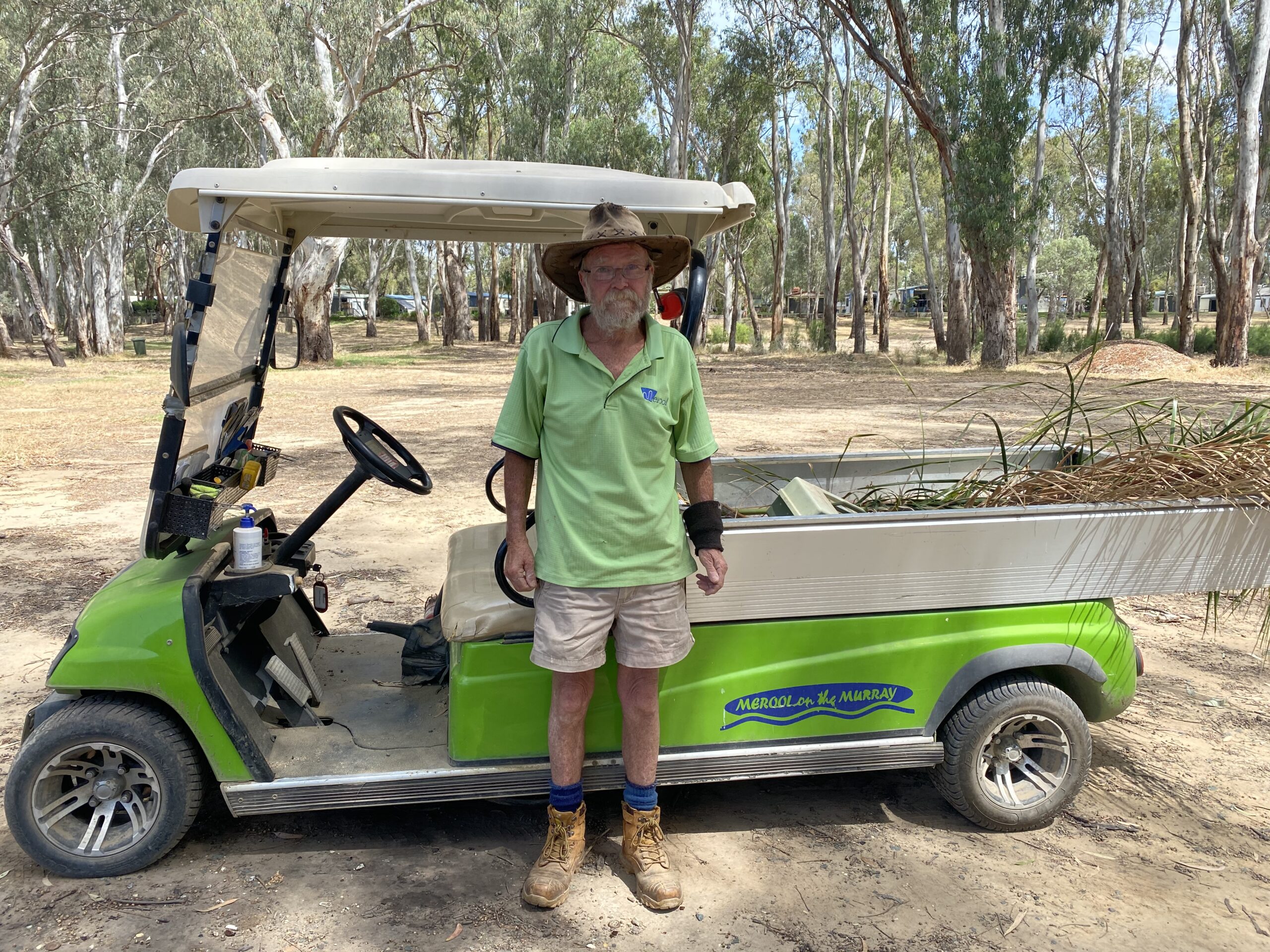 George Dick wearing his work uniform of a green t-shirt, shorts, work boots and a wide brimmed hat standing proudly infront of his green golf kart in the holiday park gardens. 