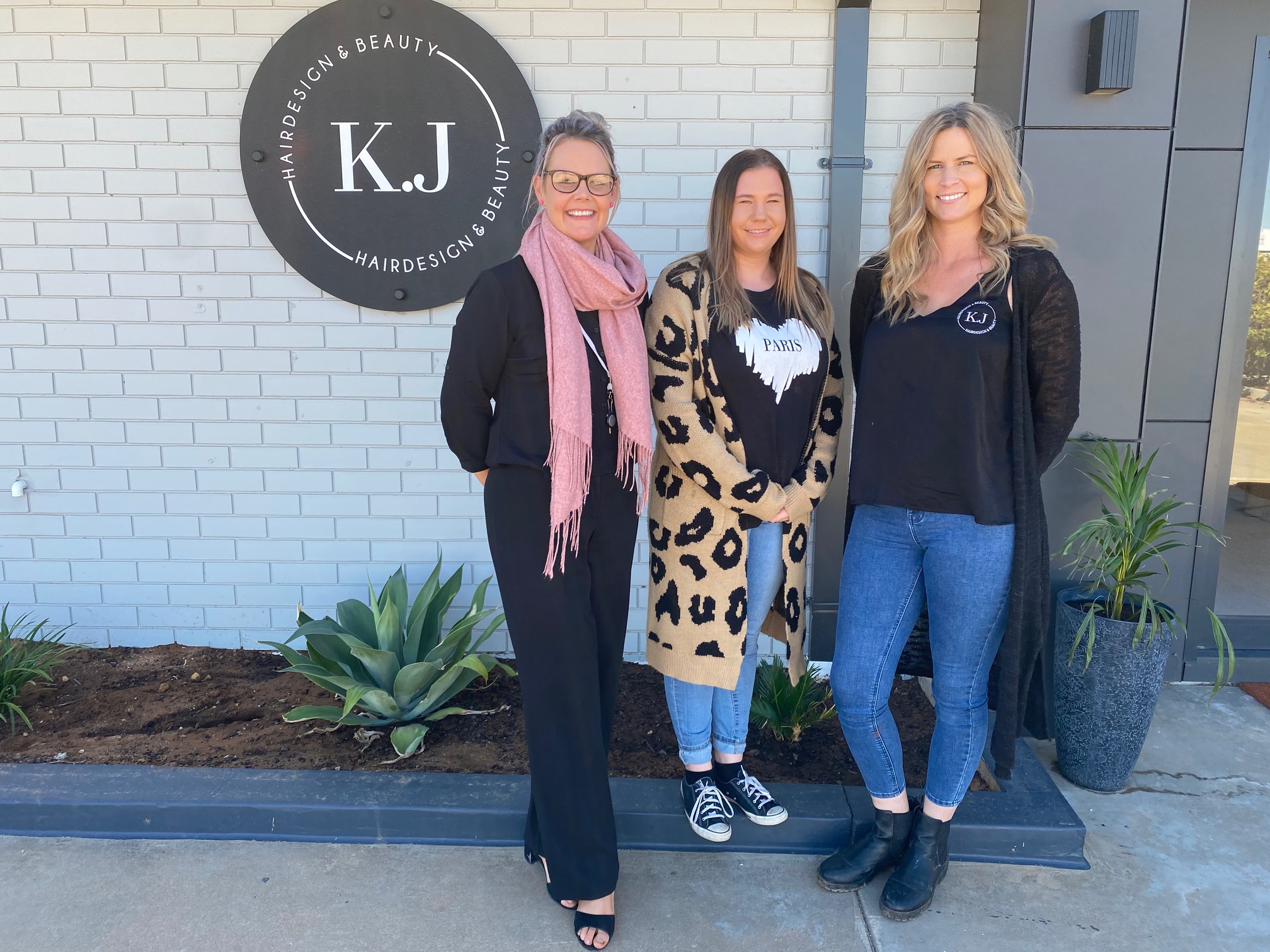 Three women standing in front of a white brick wall with the KJ Hair Designs signage